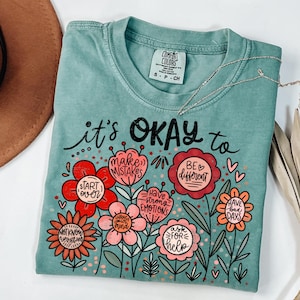 May include: A teal Comfort Colors t-shirt with the phrase "it's OKAY to" above a floral design. The flowers contain phrases like "make mistakes" and "have bad days". A silver necklace and a brown hat are partially visible.