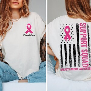 May include: Cream-colored t-shirt with a pink ribbon and the text "#TeamOlivia" on the front. The back of the shirt features a pink ribbon design, a star-studded pattern, and the words "Support Squad" and "Breast Cancer Awareness".
