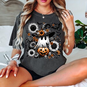 May include: Dark gray t-shirt featuring a Halloween-themed design. The graphic includes a ghost wearing a witch hat, surrounded by sunflowers, pumpkins, and bats. The design is in shades of black, white, orange, and yellow.