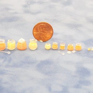 Puede incluir: Un conjunto de tarros de miel en miniatura de varios tamaños, que van desde pequeños hasta grandes, con un centavo para la escala. Los tarros están hechos de vidrio transparente y llenos de un líquido ámbar claro. El conjunto incluye un pequeño cucharón de miel de madera.