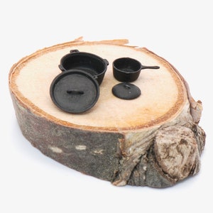 Könnte beinhalten: Miniatur-Kochgeschirr-Set aus schwarzem Gusseisen, einschließlich eines Topfes mit Deckel, einer kleinen Pfanne und einem separaten Deckel, angeordnet auf einer natürlichen Holzscheibe. Das Set ist für Dekorationszwecke oder Puppenhäuser konzipiert.