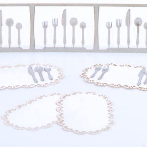 Könnte beinhalten: Ein Set aus vier weißen Spitzen-Platzsets mit silbernem Besteck, bestehend aus Gabeln, Messern und Löffeln. Das Besteck ist ordentlich auf jedem Platzset angeordnet.