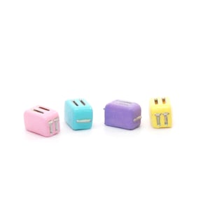 Op de afbeelding: Vier miniatuur-toasters in verschillende pastelkleuren: roze, blauw, paars en geel. Elke toaster heeft een zilveren metalen ontwerp aan de voorkant.