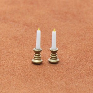 Puede incluir: Dos velas en miniatura con cera blanca y pequeñas llamas amarillas, cada una colocada en una base de candelabro de tono dorado metálico con un diseño acanalado y escalonado. Estos pequeños artículos decorativos tienen un acabado liso y forma cilíndrica.