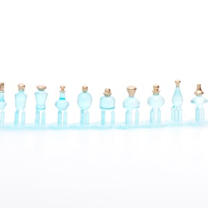 Pode incluir: Um conjunto de 12 garrafas de vidro em miniatura com tampas douradas. As garrafas estão dispostas em uma fila sobre uma base azul transparente. As garrafas são de diferentes formas e tamanhos.