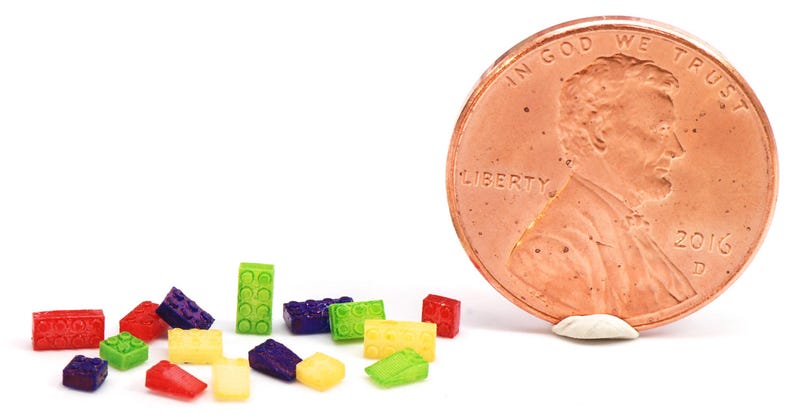 May include: A collection of colorful miniature plastic building blocks, some red, yellow, green, and purple, arranged in a row on a white surface. A copper penny coin is to the right of the blocks.