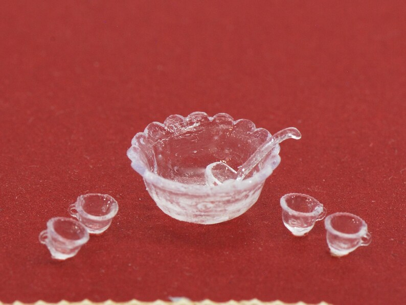 May include: A miniature clear plastic punch bowl set with a scalloped edge, a ladle, and four small cups. The set is designed for a dollhouse or miniature display. The set is on a red background.