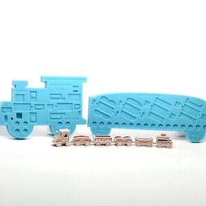 Puede incluir: Un molde de silicona azul con forma de locomotora y vagones de tren. El molde está diseñado para crear decoraciones de tren comestibles. Se muestra un conjunto de pequeños vagones de tren marrones junto al molde.