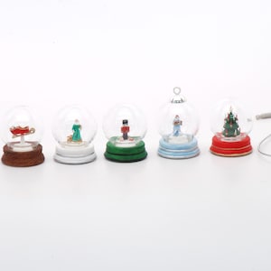 May include: Five miniature snow globes with different Christmas scenes. The snow globes are arranged in a row from left to right. The scenes include a red sled, a woman in a green dress, a nutcracker, a couple in a winter scene, and a Christmas tree.