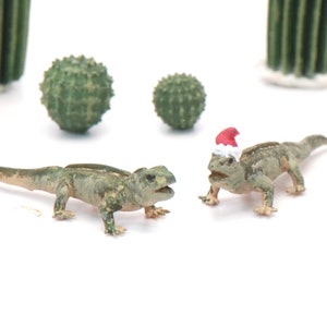 May include: Two small green lizards, one wearing a red Santa hat, on a white background.