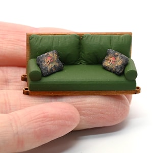 May include: Miniature green sofa with two matching pillows. The sofa has a wooden frame and armrests. The pillows are black with a floral pattern. The sofa is held in a person's hand.