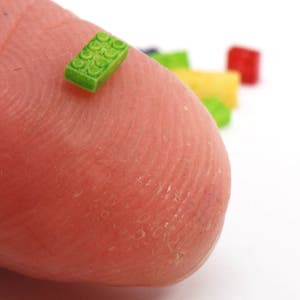 May include: A tiny green Lego brick on a fingertip, with other colorful Lego bricks in the background.