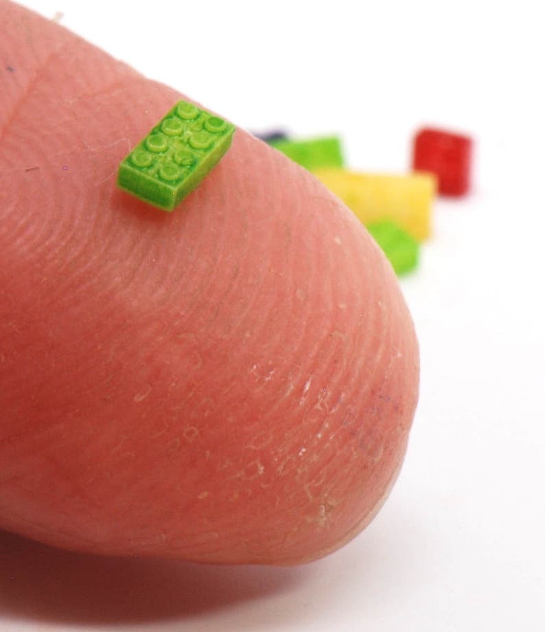 May include: A tiny green Lego brick on a fingertip, with other colorful Lego bricks in the background.