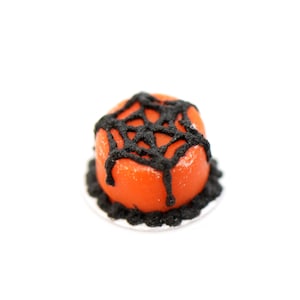 Könnte beinhalten: Ein kleiner orangefarbener Kuchen mit schwarzem Zuckerguss in Form eines Spinnennetzes.