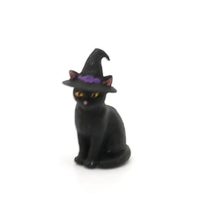 May include: A black cat figurine wearing a witch's hat with a purple band. The cat has yellow eyes and pink ears. The figurine is set against a white background, perfect for Halloween decorations.