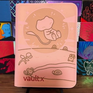 May include: Pink Vault X trading card binder featuring a cartoon illustration of a Mew character. The binder has a pink background with a circular design and the Vault X logo. Other card binder designs are visible in the background.