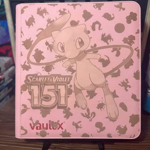Può includere: Raccoglitore di carte rosa con un personaggio dei cartoni animati Mew e la scritta "Scarlet & Violet 151". Il raccoglitore presenta un motivo ripetuto di sagome marroni di vari personaggi. Il marchio "Vaultx" è stampato in basso.