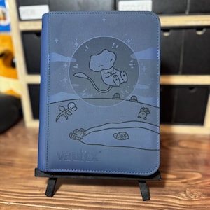 May include: A navy blue Vault X trading card binder. The cover features a cartoon illustration of a character inside a circle with a moon and stars. The binder is displayed on a small black easel.