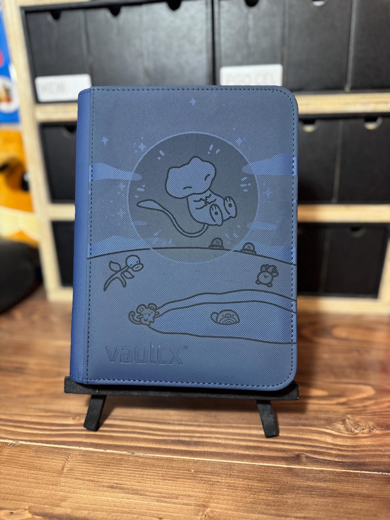 May include: A navy blue Vault X trading card binder. The cover features a cartoon illustration of a character inside a circle with a moon and stars. The binder is displayed on a small black easel.
