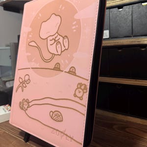 May include: A pink card binder with a black zipper and a brown illustration of a cartoon character in a circle. The binder has a brown illustration of a landscape with cartoon animals and the word "Zovly".