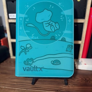 May include: A teal Vault X trading card binder featuring a cartoon illustration of a character. The binder has a stitched border and the Vault X logo. The background includes a cartoon landscape with additional character illustrations.