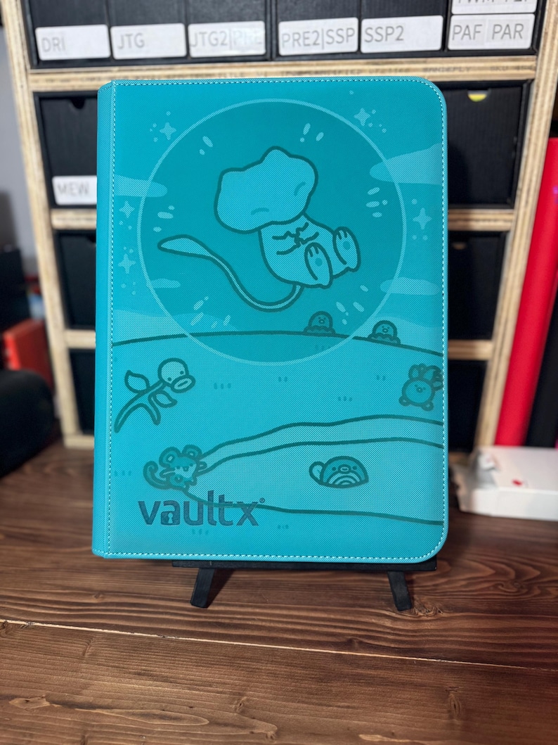 May include: A teal Vault X trading card binder featuring a cartoon illustration of a character. The binder has a stitched border and the Vault X logo. The background includes a cartoon landscape with additional character illustrations.