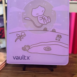 May include: A lavender Vault X binder featuring a cartoon illustration of a character in a circle with a moon and clouds. The binder has a brown line drawing of a landscape with other cartoon characters. The Vault X logo is at the bottom.