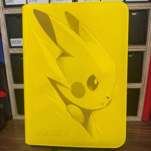 May include: A yellow Vault X trading card binder with a Pikachu design. The Pikachu graphic is a light yellow, and the binder has a black stand. The word "vaultx" is printed on the bottom of the binder.