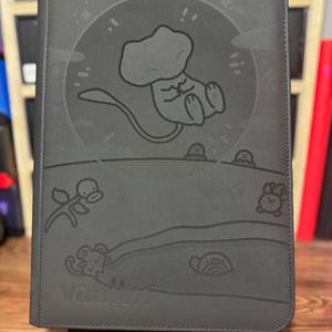 May include: A dark gray binder with a cartoon illustration of a creature in a circle, with other cartoon figures below. The word "Vault X" is embossed at the bottom. The binder is standing on a black stand.