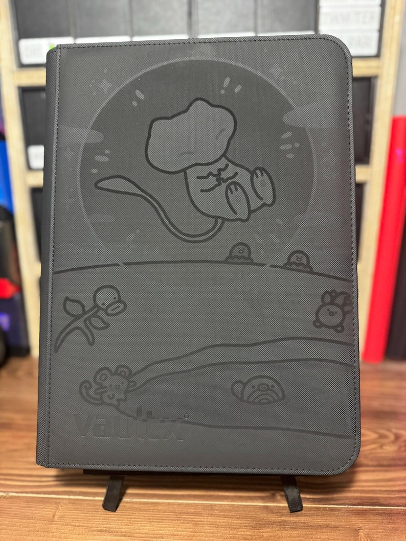 May include: A dark gray binder with a cartoon illustration of a creature in a circle, with other cartoon figures below. The word "Vault X" is embossed at the bottom. The binder is standing on a black stand.
