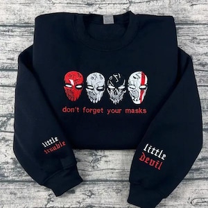 Może przedstawiać: Czarna bluza z czterema stylizowanymi wzorami masek w kolorze czerwonym i białym. Pod maskami znajduje się napis "don't forget your masks". Lewy rękaw ma napis "little trouble", a prawy "little Devil".