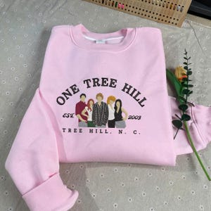 May include: A light pink sweatshirt with the text "ONE TREE HILL" in black arched lettering. Below the text is a graphic of five people and the text "EST 2003 TREE HILL, N. C."