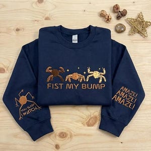 Puede incluir: Una sudadera azul marino de cuello redondo con figuras de cangrejo bordadas en marrón y el texto FIST MY BUMP en el pecho. La manga izquierda muestra un gráfico de cangrejo con el texto YOU SLEEP I WATCH y la derecha el texto AMAZE! AMAZE! AMAZE!