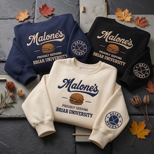 Op de afbeelding: Drie crewneck sweatshirts in marineblauw, zwart en crème. Elk heeft de tekst "Malone's" boven een burgerafbeelding, met "Proudly Serving Briar University" eronder. Een Briar Hockey-logo staat op de linkermouw.