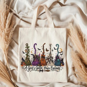 May include: A cream-colored canvas tote bag featuring a graphic print of five stylized witch brooms in green, purple, orange, white, and blue. The design includes small bats, stars, and the text A Girl's Gotta Have Options in black script.