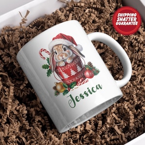 May include: White ceramic mug featuring a Christmas-themed design. The mug showcases a bunny wearing a Santa hat, nestled inside a red cup with Christmas tree patterns. The name "Jessica" is written in green script.