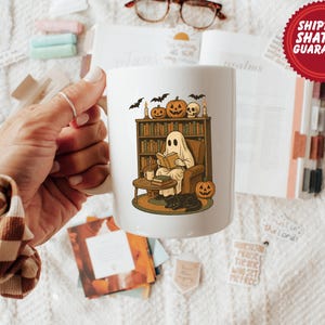 May include: White ceramic mug featuring a Halloween-themed illustration of a ghost reading in a cozy library setting. The design includes pumpkins, bats, a skull, and a black cat. A perfect gift for book lovers and Halloween enthusiasts.