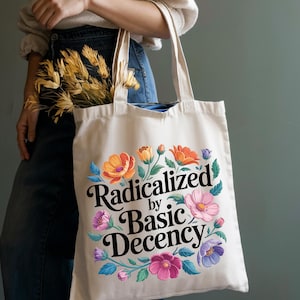 May include: Cream-colored tote bag with the phrase "Radicalized by Basic Decency" in black script, surrounded by colorful flowers in orange, pink, and purple. The bag is held by a person.