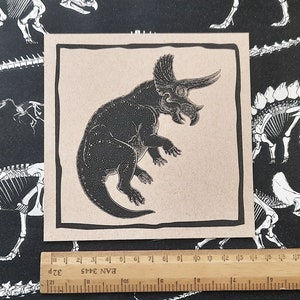 May include: A black and white illustration of a triceratops dinosaur on a beige background. The dinosaur is facing right and has a large frill on its head. The illustration is framed with a black border.