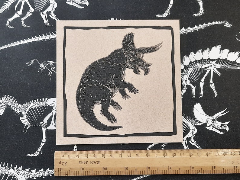 May include: A black and white illustration of a triceratops dinosaur on a beige background. The dinosaur is facing right and has a large frill on its head. The illustration is framed with a black border.