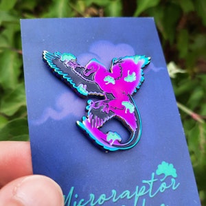 May include: A colorful enamel pin featuring a Microraptor dinosaur. The pin has a black body with iridescent wings and a vibrant pink and teal design. The pin is on a blue card with the word "Microraptor" in cursive.