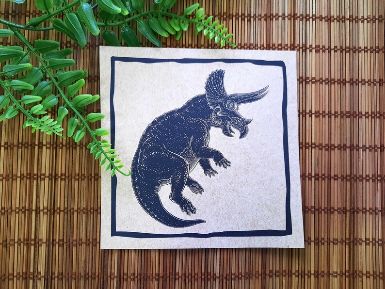 May include: A black and white illustration of a Triceratops dinosaur on a beige background with a black border.