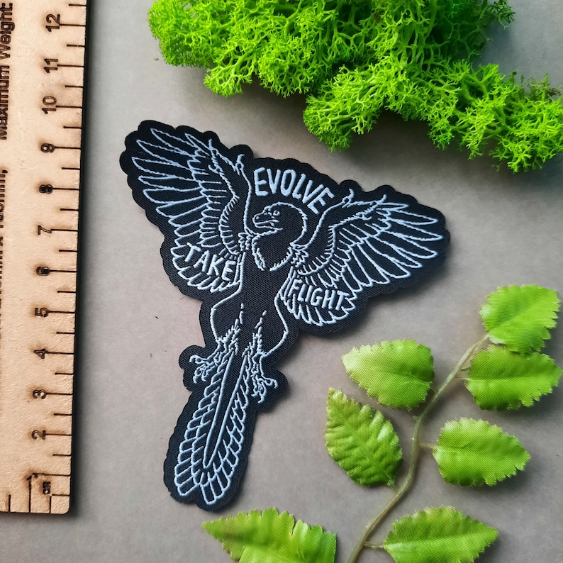 May include: Black embroidered patch with a white outline of a bird in flight. The bird has the words "Evolve" and "Take Flight" written on it.
