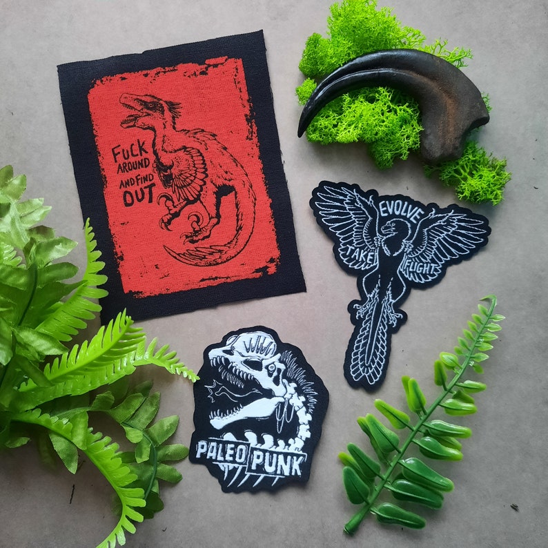 May include: Three iron-on patches featuring dinosaur and bird designs. The first patch is red with black text that reads "Fuck Around and Find Out" and a black silhouette of a dinosaur. The second patch is black with white text that reads "Evolve Take Flight" and a white silhouette of a bird. The third patch is black with white text that reads "Paleo Punk" and a white silhouette of a dinosaur head.