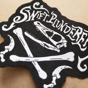 May include: Black patch with white embroidered design. The patch features the text "Swift Plunderer" in a stylized font, along with a dinosaur skull and crossed bones. The patch has a black background and a white border.