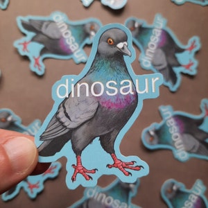 May include: A blue sticker with a cartoon pigeon on it. The pigeon is gray and has a white ring around its eye. The sticker says "dinosaur" in white letters.