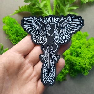 May include: Black iron-on patch with white embroidery of a bird with outstretched wings. The text "Evolve Take Flight" is embroidered around the bird.