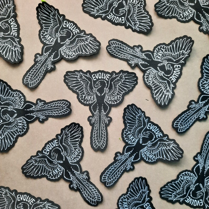 May include: Multiple black iron-on patches with white embroidery. Each patch features a stylised bird with outstretched wings and the text "EVOLVE TAKE FLIGHT".