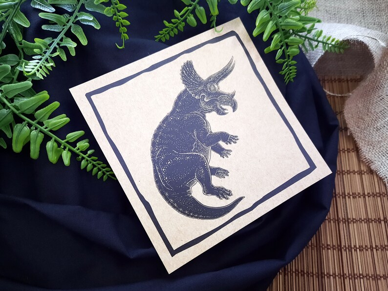 May include: A black and white illustration of a triceratops dinosaur on a brown card with a black border.
