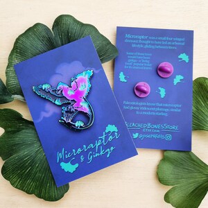 May include: A vibrant enamel pin featuring a Microraptor dinosaur with iridescent pink and blue coloring, set against a dark blue background. The pin is displayed with a matching backing card and two purple pin backs. The card reads "Microraptor & Ginkgo."